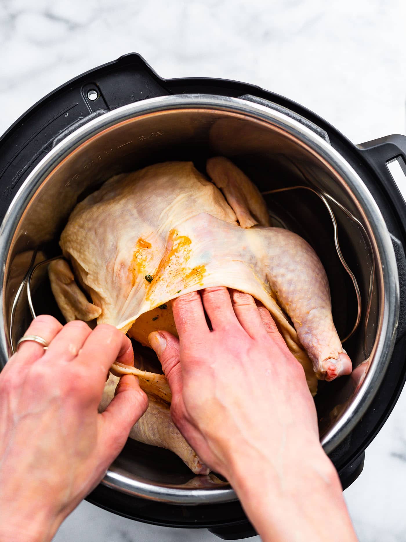 a whole chicken in an instant pot with a woman's hands pulling the skin from the meat and spreading a marinade onto the meat by the legs