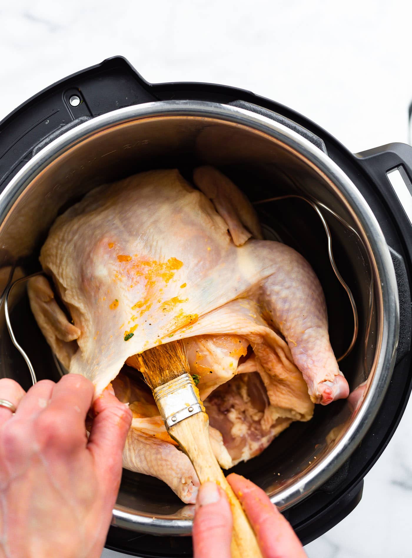 a whole chicken in an instant pot with a woman's hands holding the skin from the meat and using a brush to brush marinade onto the meat near the legs of the chicken