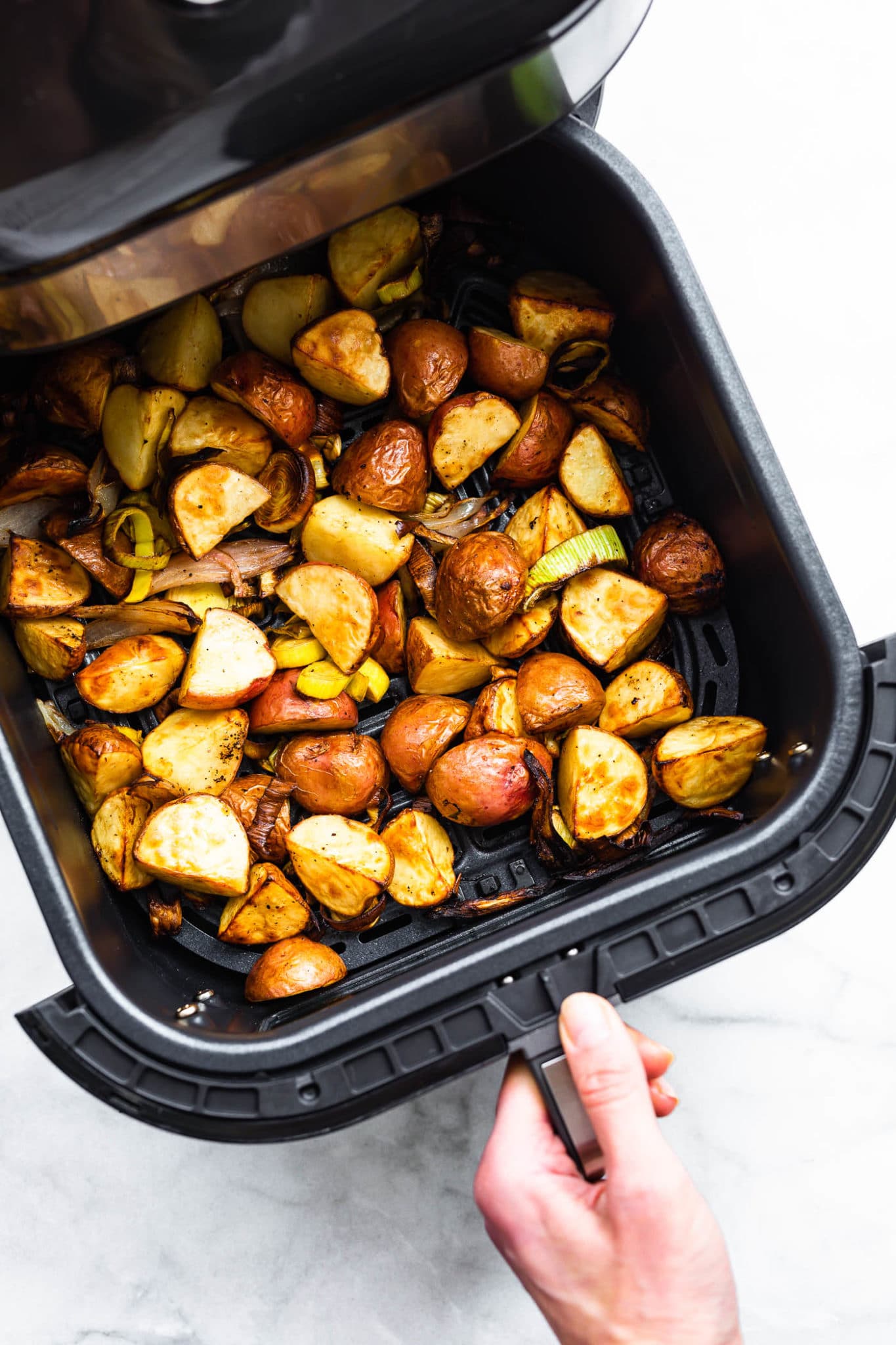 a hand pulling the basket out of an air fryer basket full of roasted leeks and potatoes with onions