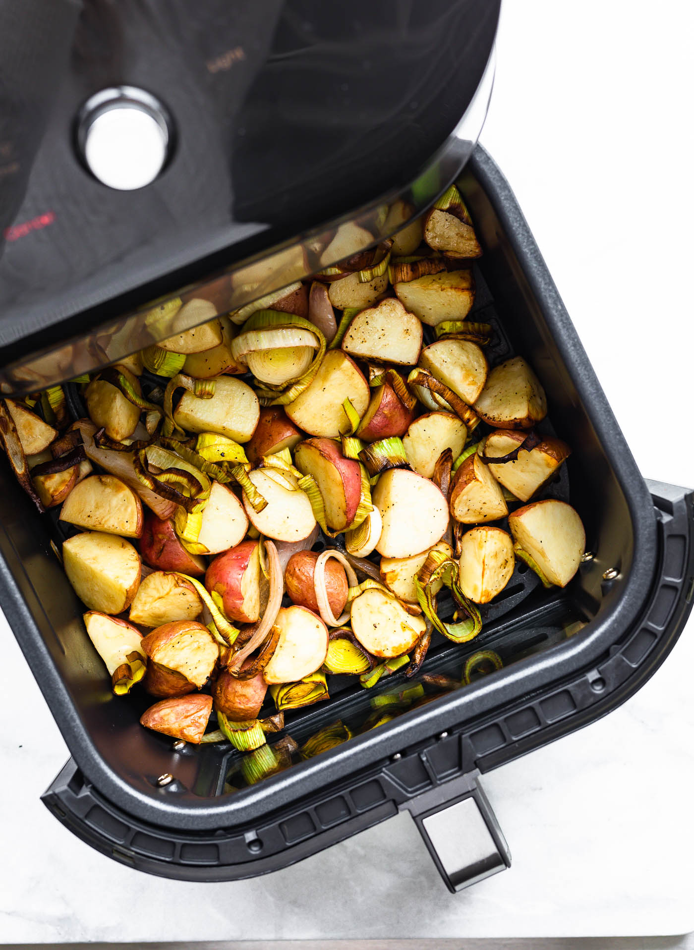 an air fryer basket sticking out of the air fryer full of roasted leeks and potatoes with onions