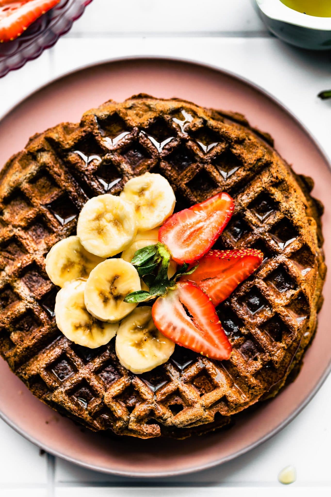 Close up overhead view one large round Vegan Buckwheat Banana Waffle on pink plate topped with strawberry and banana slices.