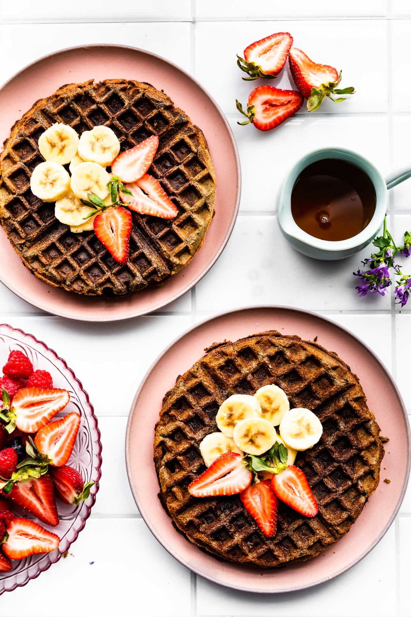 overhead view vegan buckwheat banana waffles on two pink plates topped with fresh strawberry and banana slices with a small pitcher of syrup on the side