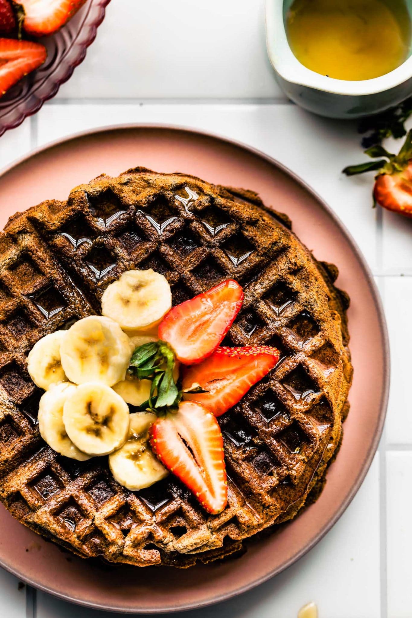 overhead image of a cooked vegan buckwheat banana waffle topped with sliced bananas, sliced strawberries, and syrup