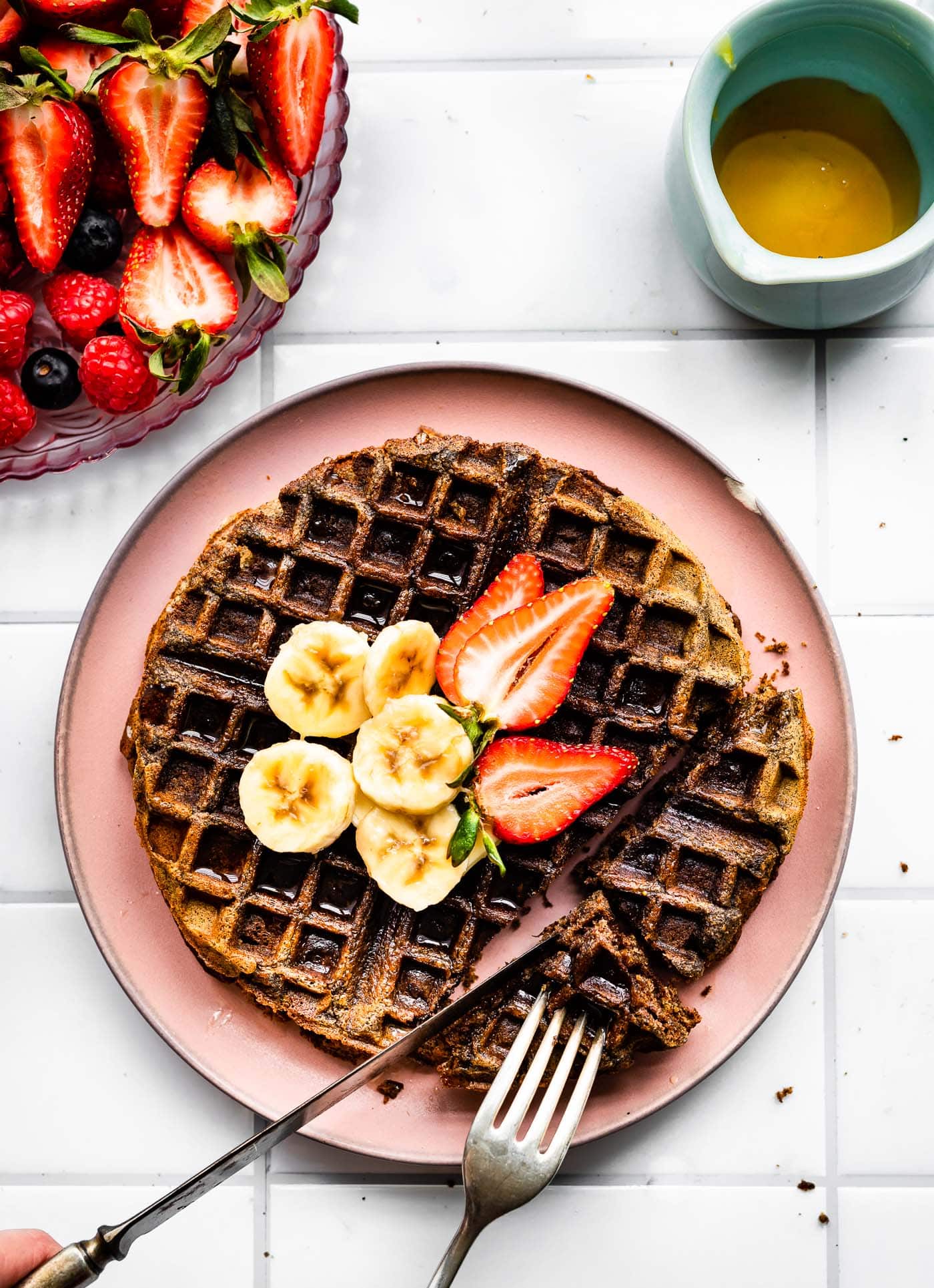 overhead image of a vegan buckwheat banana waffle on a pink plate topped with strawberries and bananas being sliced into with a knife and fork, a bowl of strawberries and blueberries, and a container of syrup