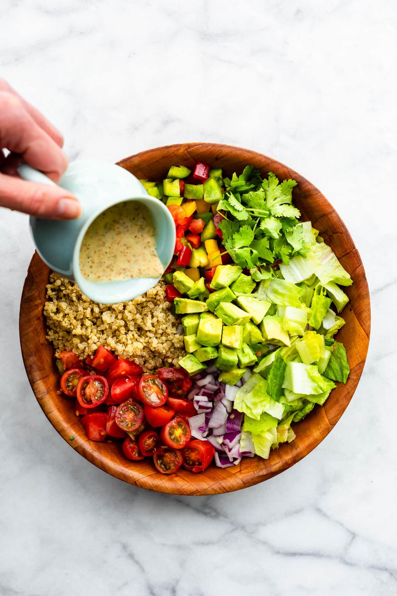 a bowl of ingredients for fiesta quinoa salad including quinoa, bell peppers, cilantro, avocado, romaine, red onion, and tomatoes with a hand pouring spicy dressing on top