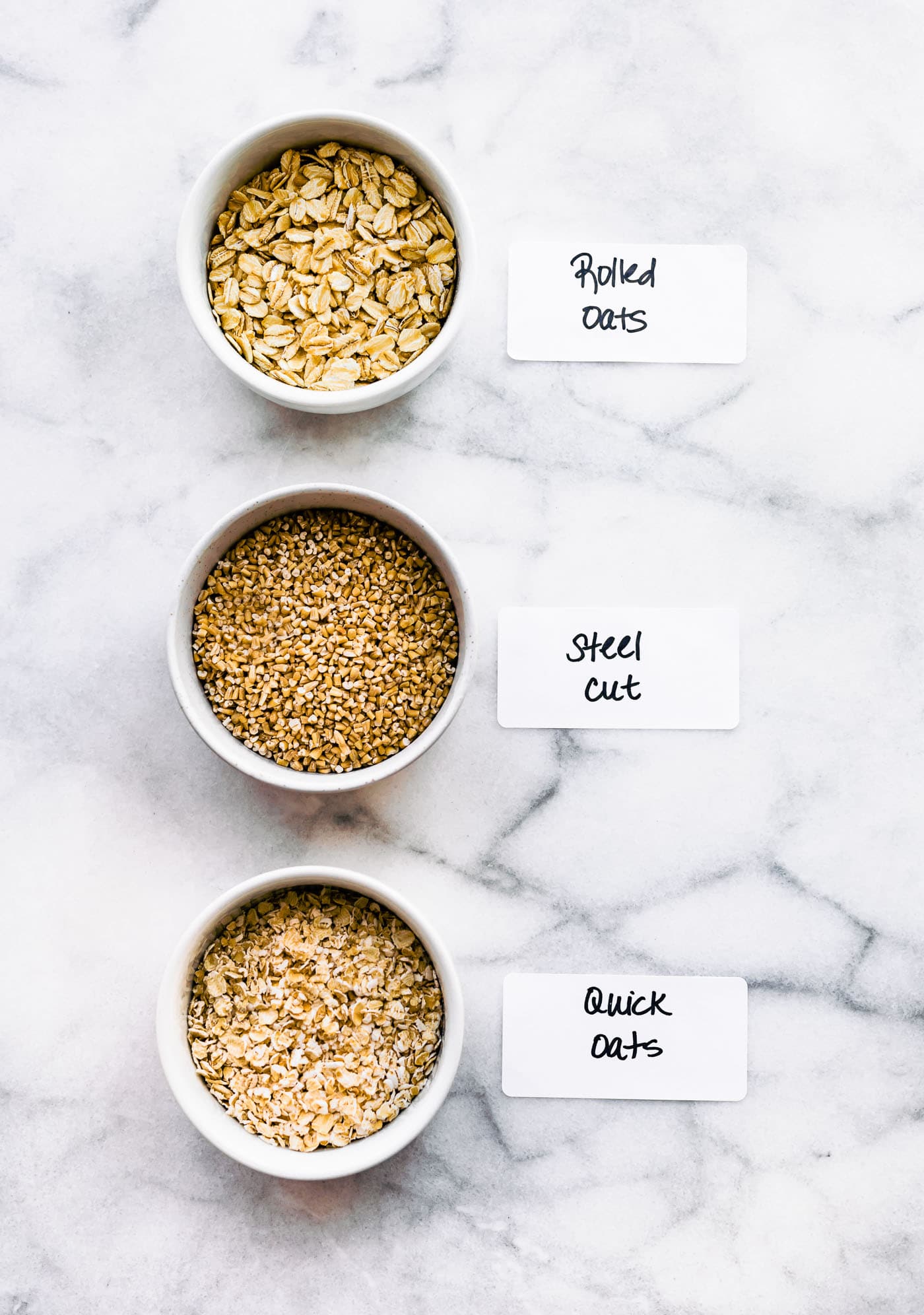 overhead image of three white bowls on a marble counter filled with different oats labeled rolled oats, steel cut, and quick oats