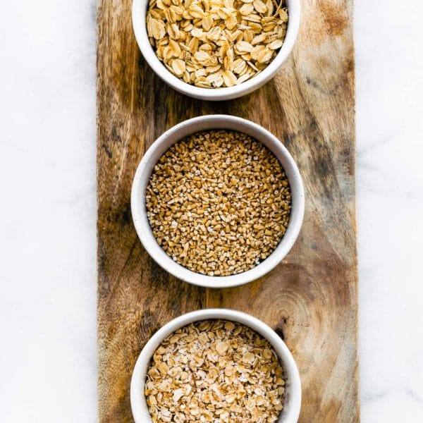 overhead image of three white bowls filled with rolled oats, quick oats, and steel cut oats on a wooden board