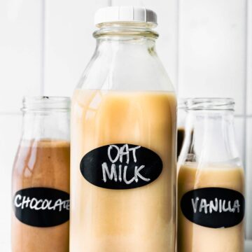 three glass jars of oat milk, the largest in the center labeled oat milk, the smaller jar on the left labeled chocolate, and the smaller. jar on the right labeled vanilla