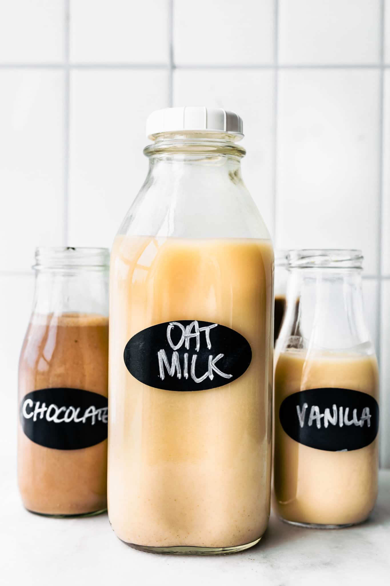 three glass jars of oat milk, the largest in the center labeled oat milk, the smaller jar on the left labeled chocolate, and the smaller. jar on the right labeled vanilla