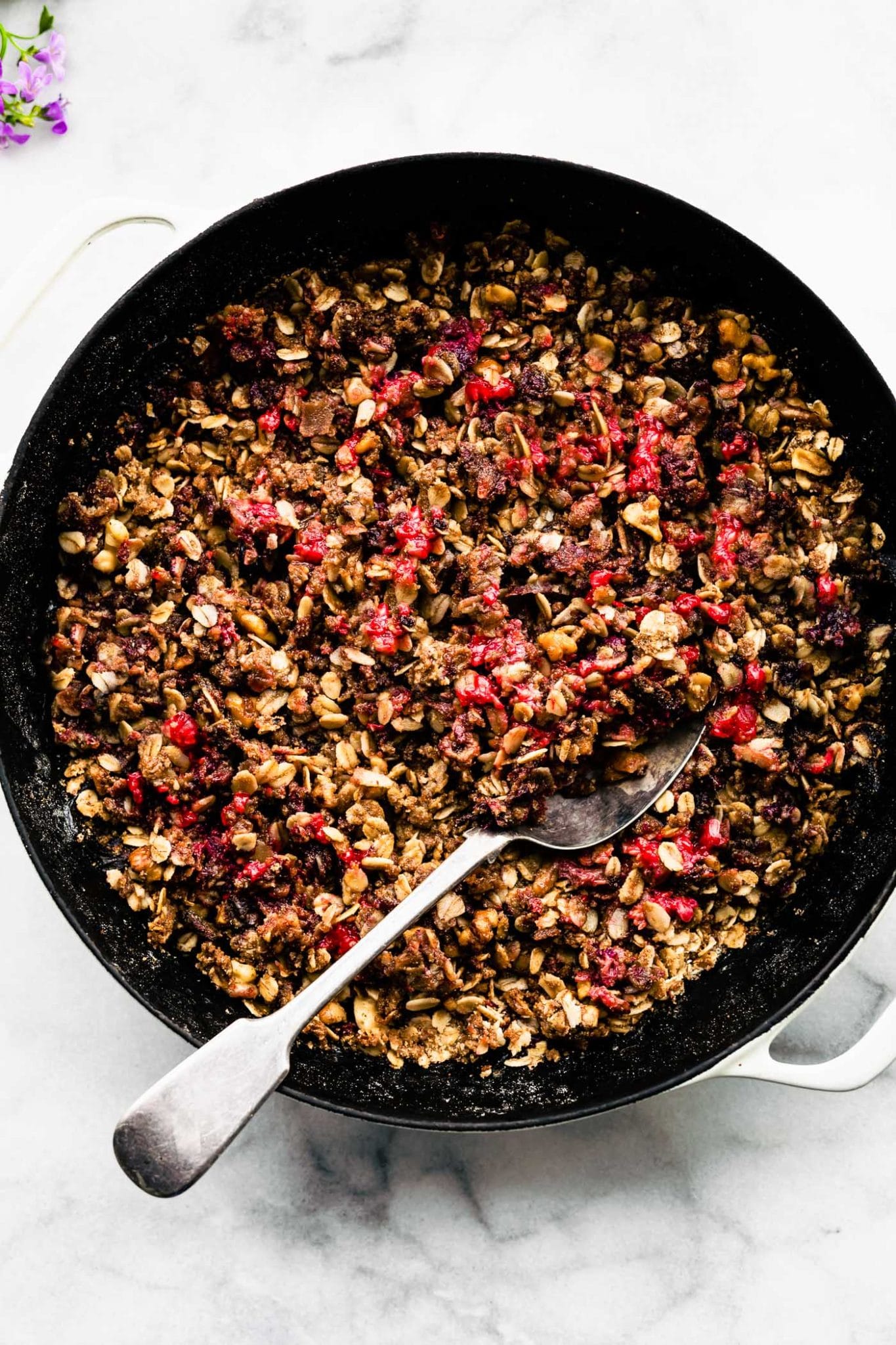 Overhead view vegan berry rhubarb crumble in skillet with large serving spoon in crumble.