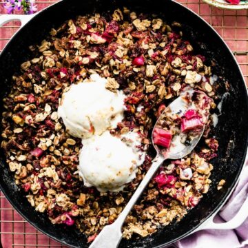 Overhead view berry rhubarb crumble in skillet with two scoops vanilla ice cream melting into top.