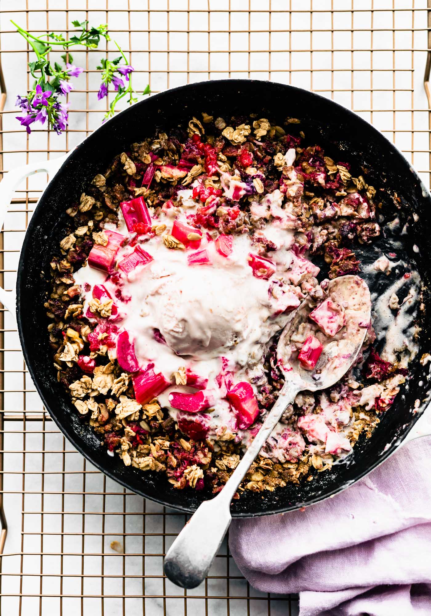 a skillet filled with berry rhubarb crumble, topped with melted ice cream and fruit. Spoon in the middle.