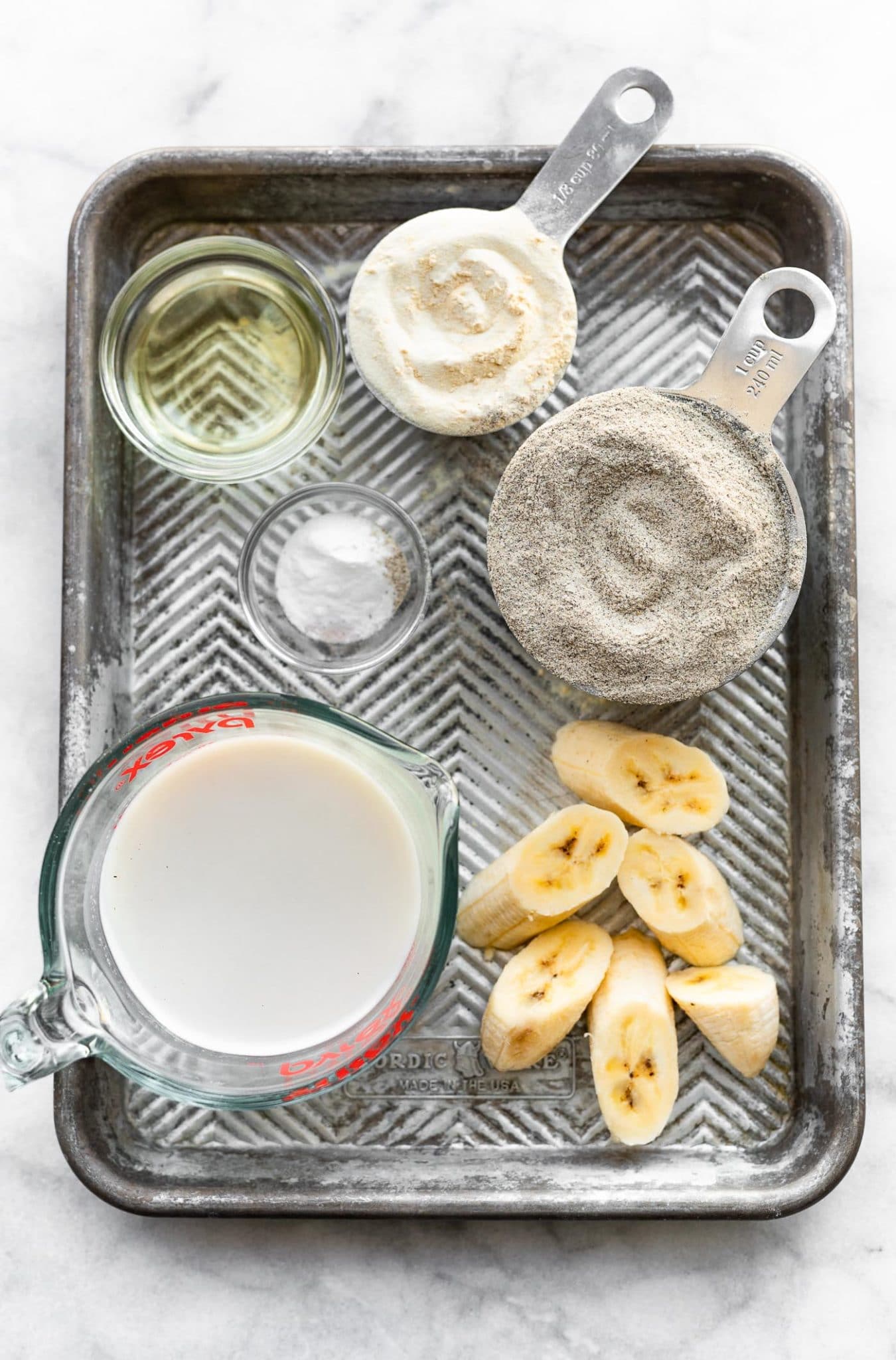 overhead image of non-dairy milk, sliced bananas, buckwheat flour, and other ingredients on a baking sheet to make vegan buckwheat banana waffles