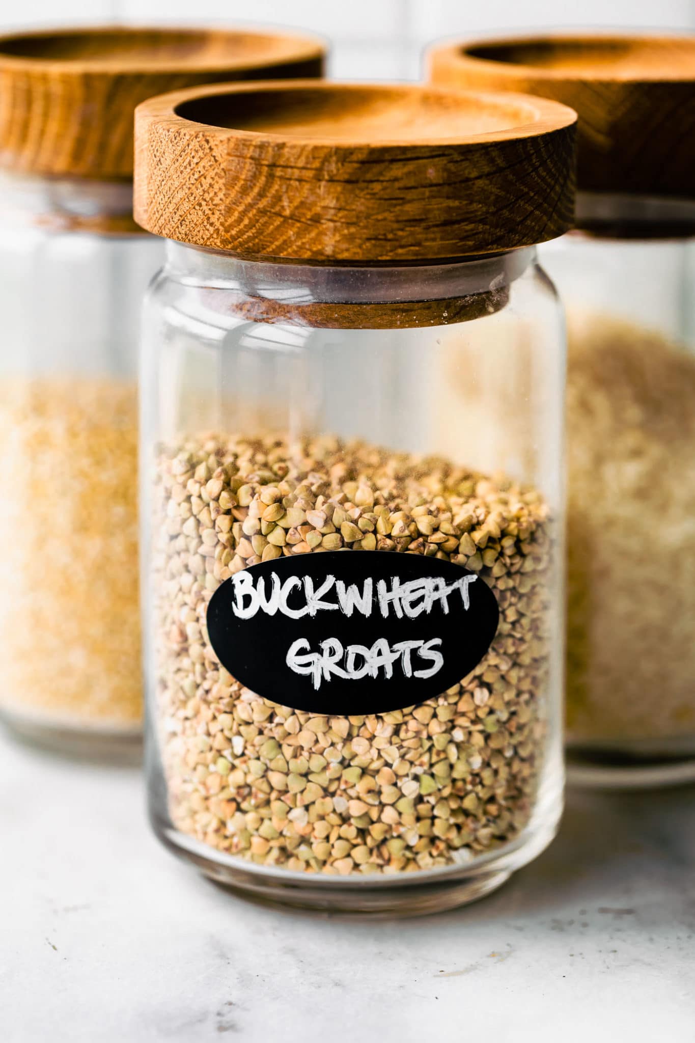 three glass jars filled with gluten free whole grains, the first labeled "buckwheat groats"