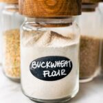 three glass jars with wooden lids full of whole grains - the first labeled "buckwheat flour"