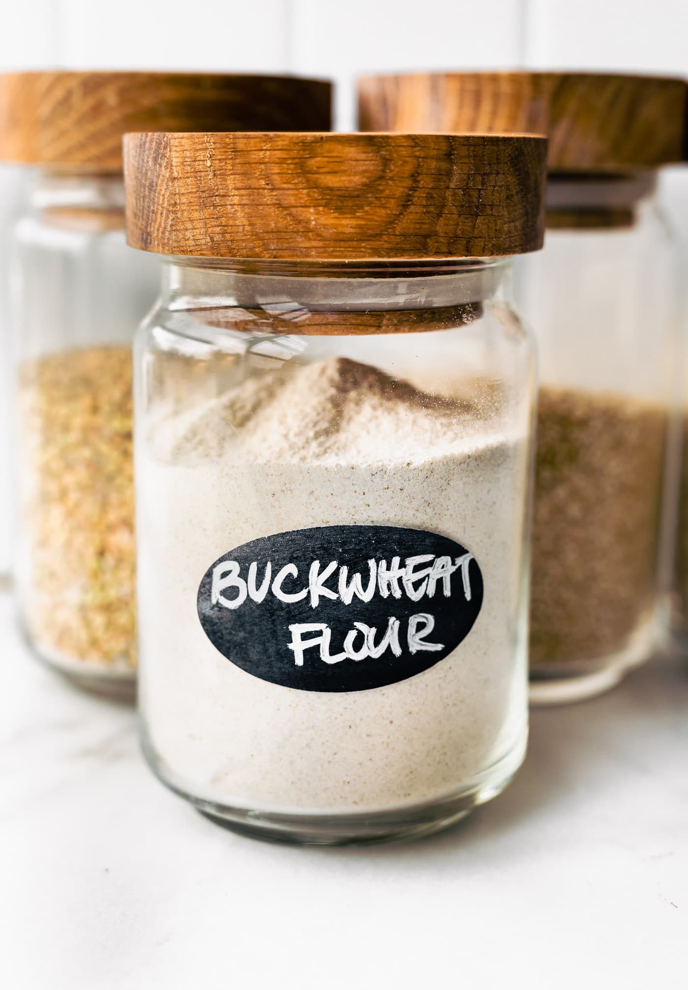 three glass jars with wooden lids full of whole grains - the first labeled "buckwheat flour"
