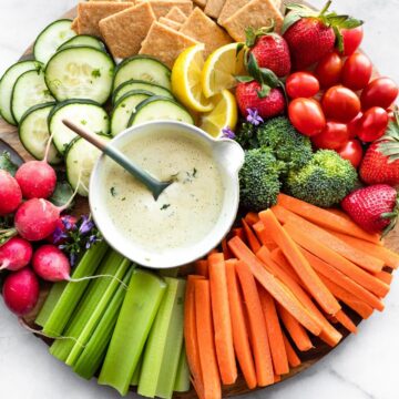 a snack platter with celery sticks, carrot sticks, broccoli florets, strawberries, grape tomatoes, gluten free crackers, lemon wedges, cucumber slices, radishes, and a small bowl of vegan ranch dressing with a spoon in it in the center