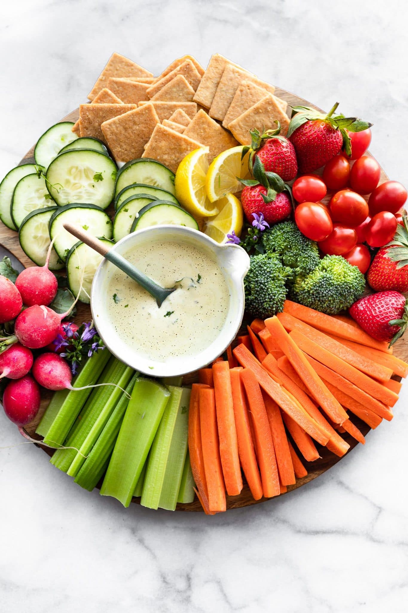 a snack platter with celery sticks, carrot sticks, broccoli florets, strawberries, grape tomatoes, gluten free crackers, lemon wedges, cucumber slices, radishes, and a small bowl of vegan ranch dressing with a spoon in it in the center