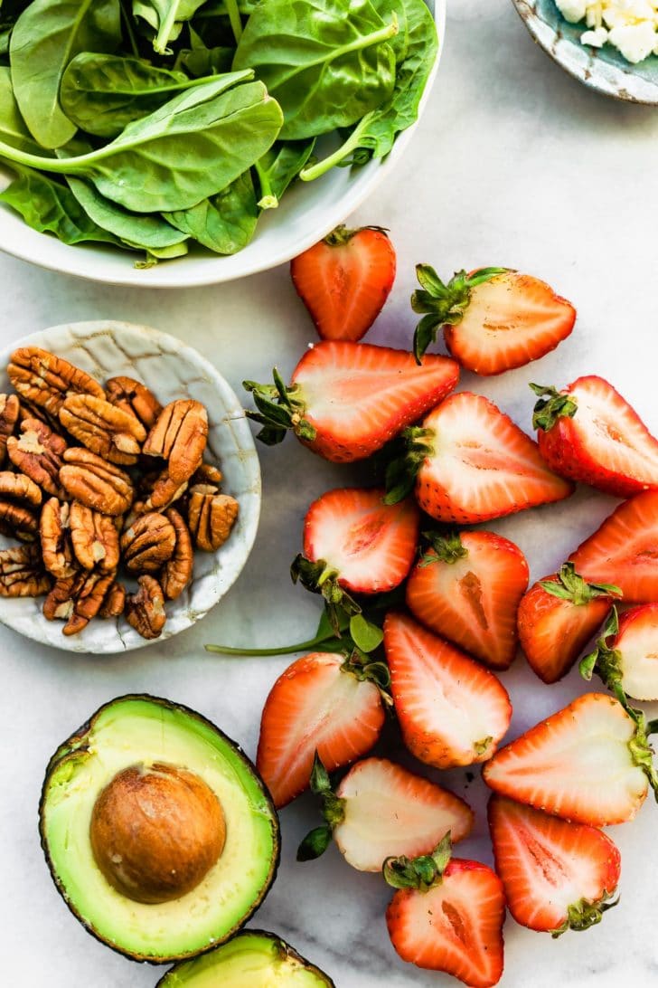 overhead image of a bowl of spinach, a small bow of feta cheese crumbles, a small bowl of pecans, halved strawberries, and a halved avocado ready to make a summer strawberry salad recipe