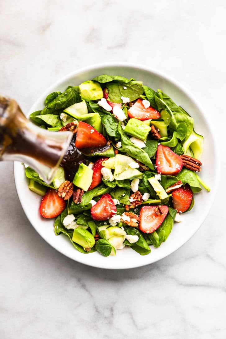 overhead image of a vinaigrette being poured over a white bowl full of summer strawberry salad