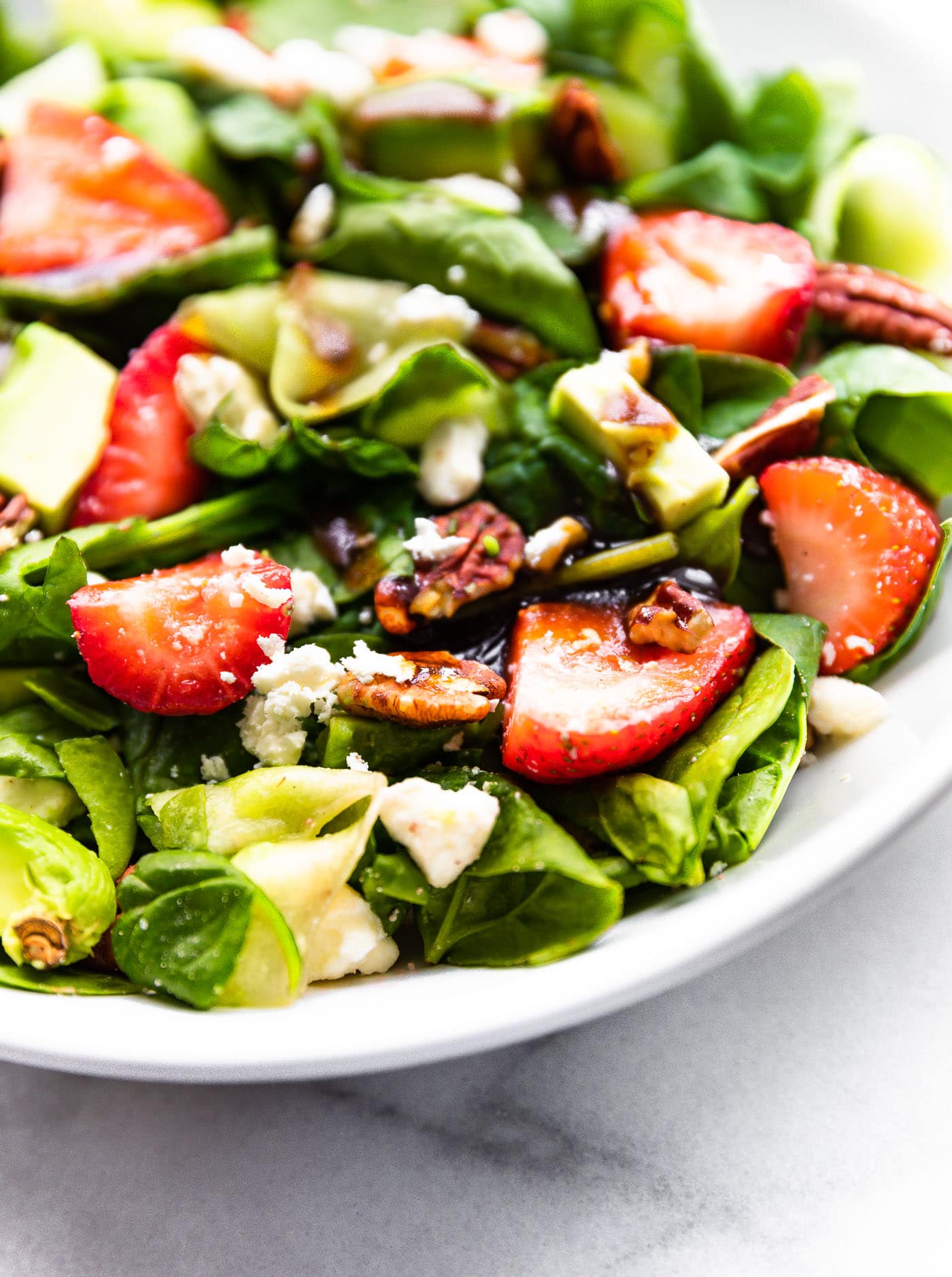close up image of a summer strawberry salad recipe with spinach, strawberries, feta, avocado, and pecans in a white bowl