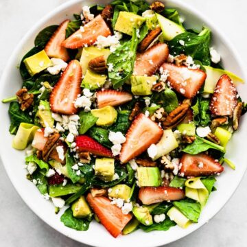 overhead image of a summer strawberry salad recipe including spinach, strawberries, avocado, feta, and pecans in a white bowl