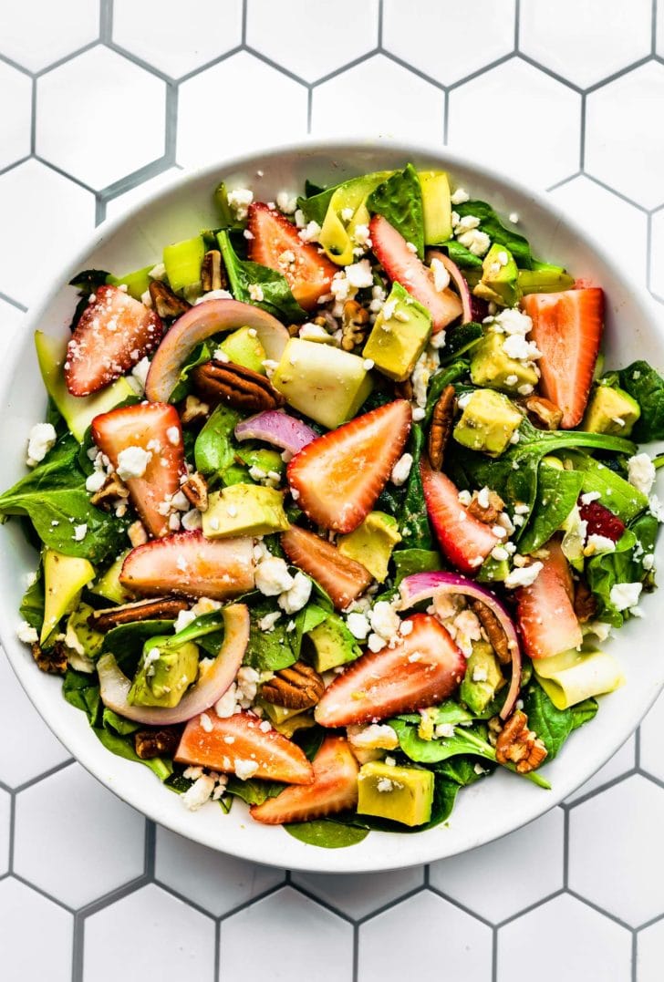 overhead image of a summer strawberry salad recipe in a white bowl including spinach, red onion, feta, avocado, strawberries, and pecans