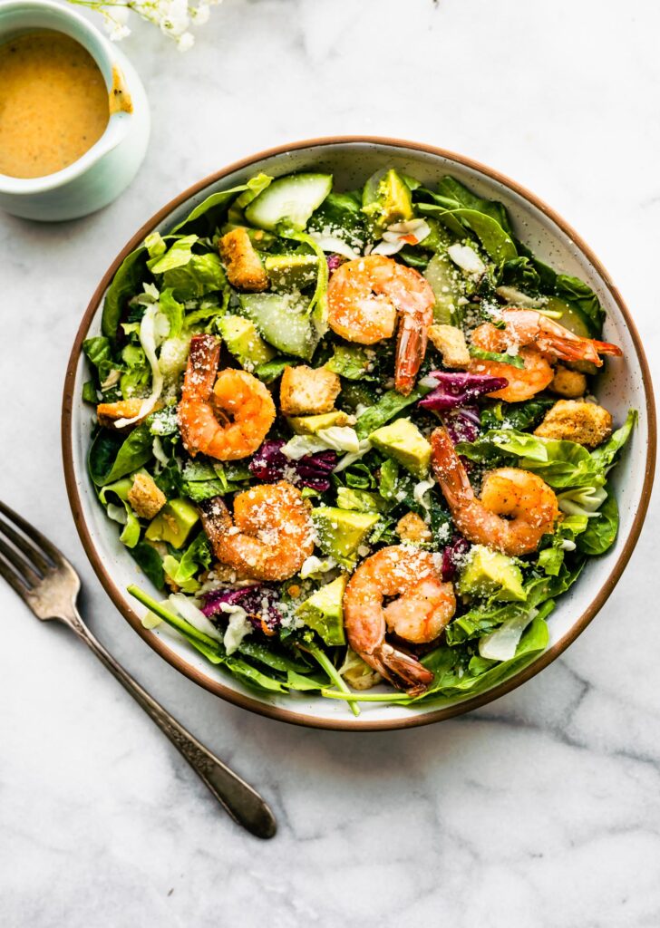 Overhead view spicy shrimp Caesar salad in white and brown bowl topped with croutons and avocado pieces.