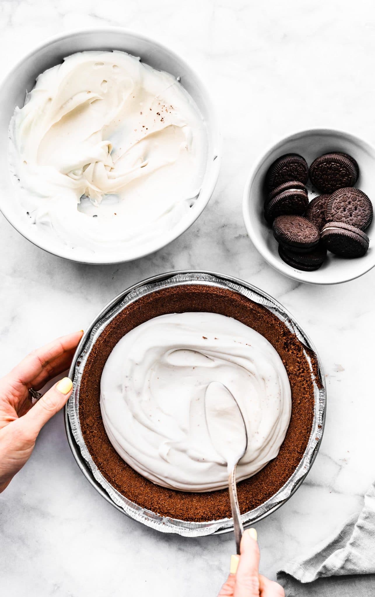 a bowl of coconut whipped cream, a bowl of chocolate creme cookies, and a pie pan full of chocolate crust with two hands using a spoon to spread a pudding and cream mixture into the center