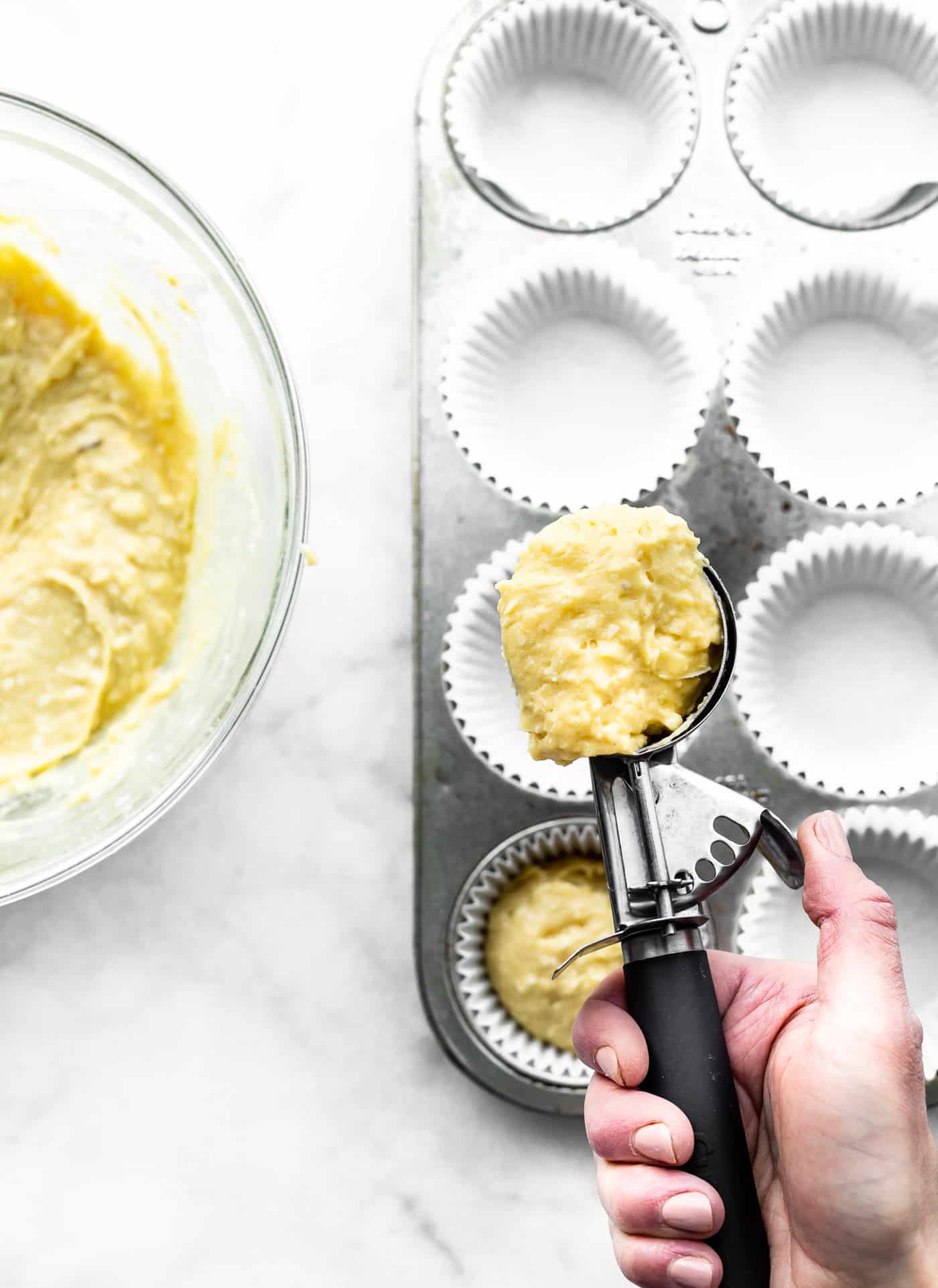 a muffin pan with paper liners, one filled with batter and a hand getting ready to fill a second cup with orange coconut oatmeal muffins batter