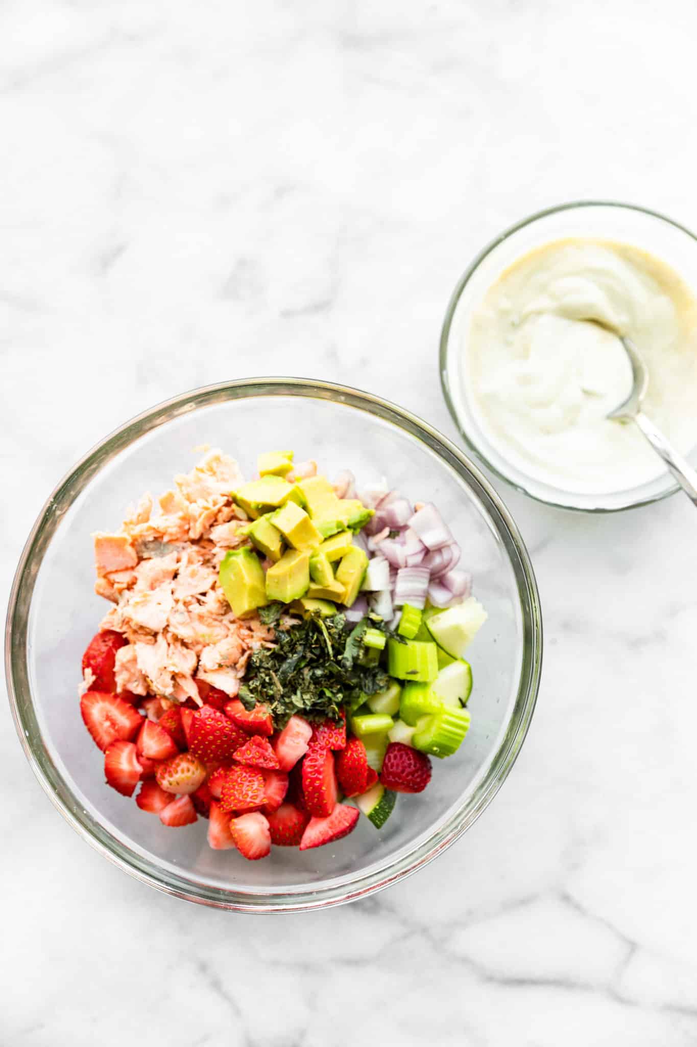 a glass bowl full of seasonings, flakes salmon, chopped strawberries, chopped celery, red onion, and avocado with a glass bowl of yogurt dressing on the side