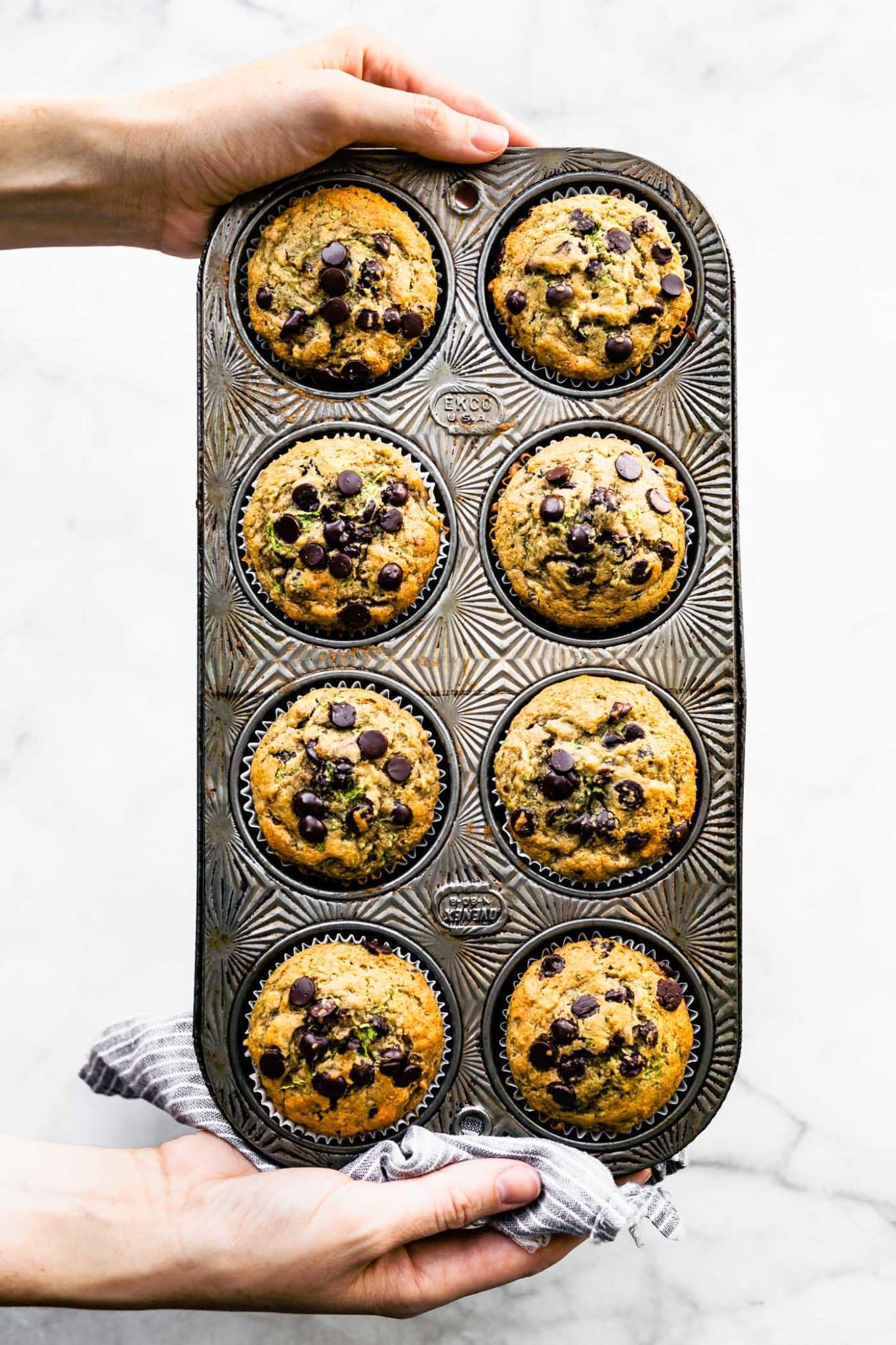 two hands holding a muffin tin with 8 baked gluten free zucchini muffins topped with chocolate chips