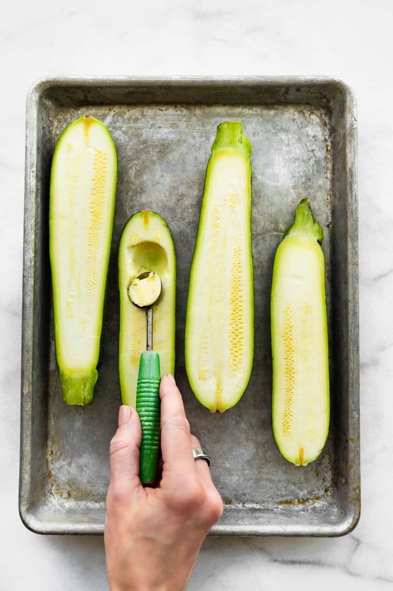 two halved zucchinis in a baking dish with a woman using a melon baller to scoop out the center of one