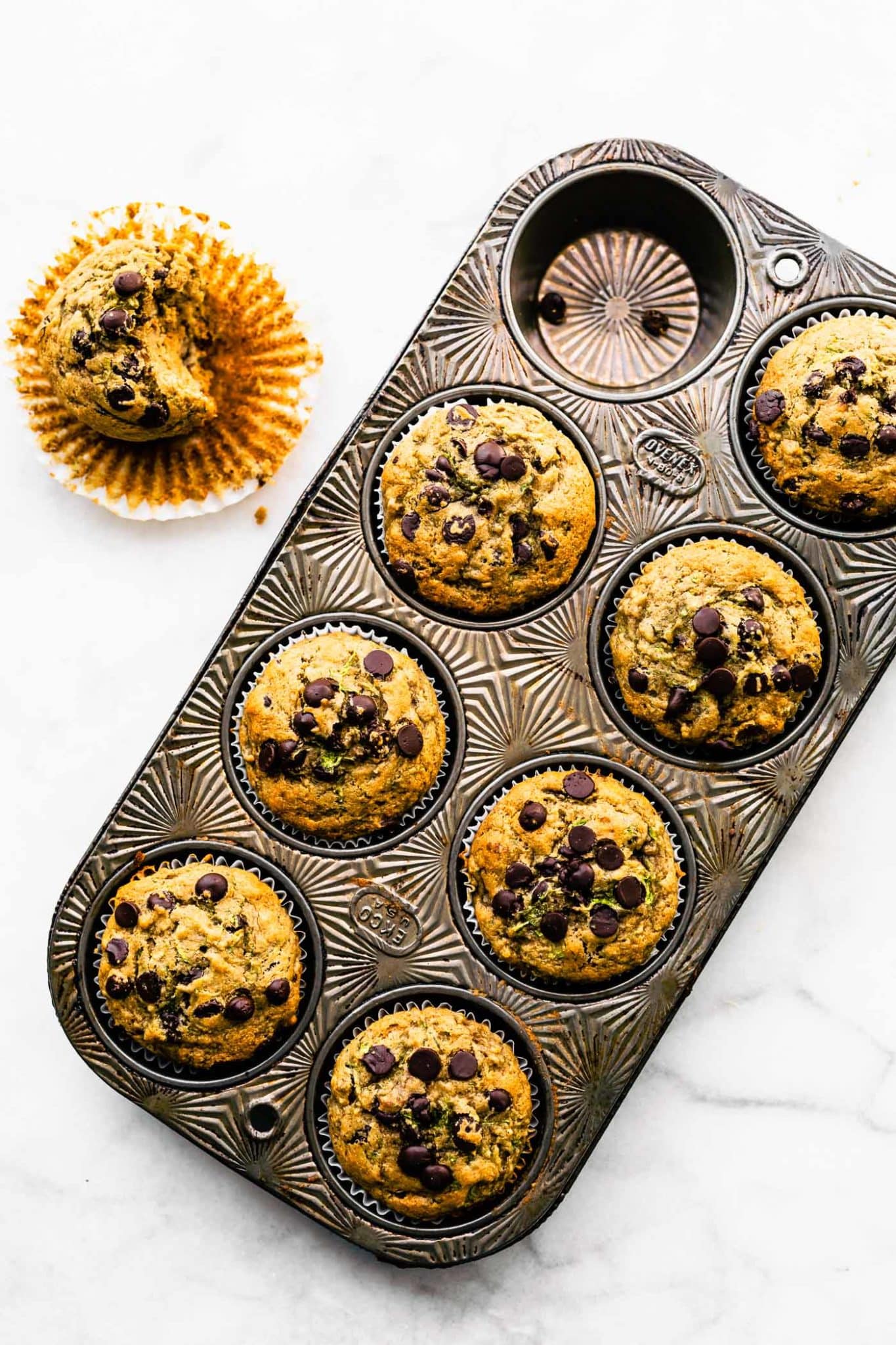 a muffin pan full of 7 baked gluten free zucchini muffins and a single muffin on the side with the liner pulled down and a bite taken out