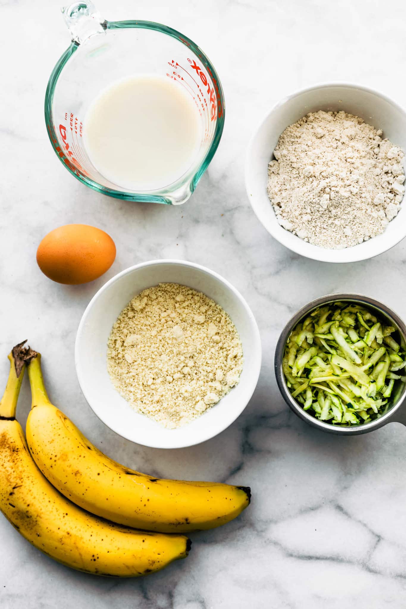 overhead image of ingredients for gluten free zucchini muffins including non-dairy milk, oat flour, almond flour, shredded zucchini, an egg, and two ripe bananas