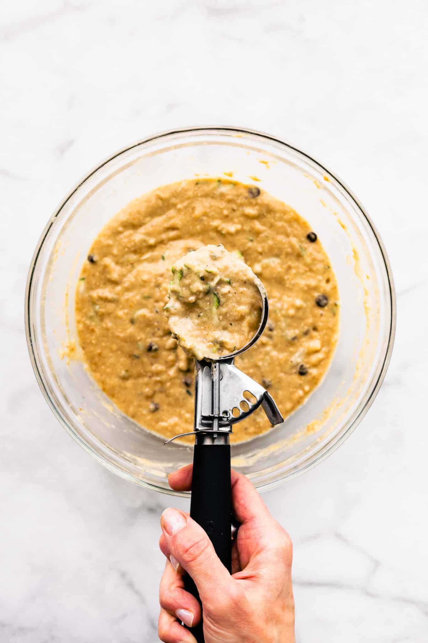 a woman's hand holding a cookie scoop full of batter over a glass bowl of gluten free zucchini muffin batter