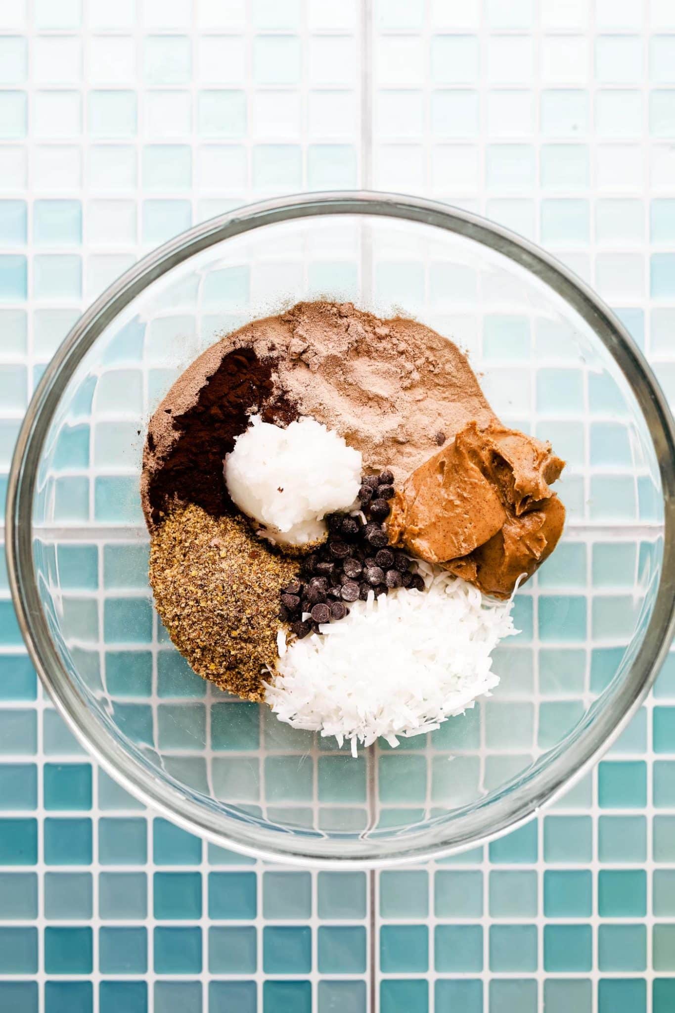 cocoa powder, protein powder, flax meal, nut butter, coconut oil, chocolate chips, and coconut flakes in a glass bowl