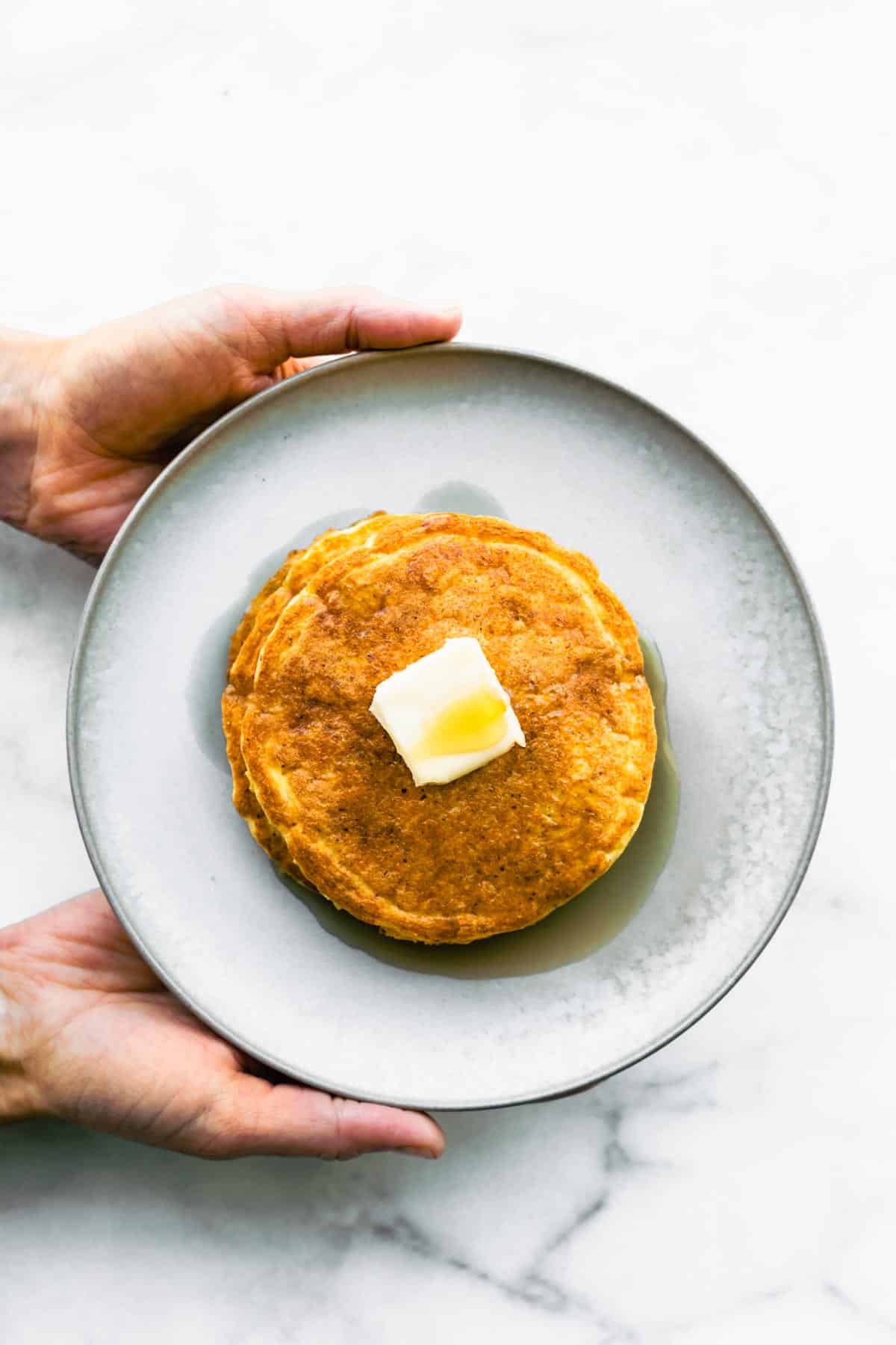 overhead image of two hands holding a plate with coconut flour pancakes topped with butter and syrup