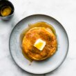 overhead image of a white plate with a stack of coconut flour pancakes topped with a pad of butter and maple syrup