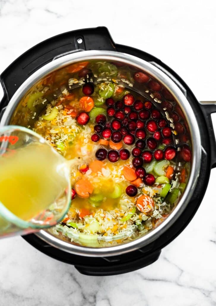 broth being poured into an instant pot with veggies, herbs, and cranberries