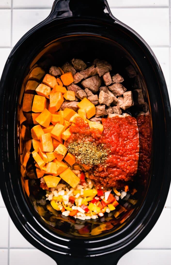 overhead image of a slow cooker with stew beef pieces, sweet potato cubes, salsa, and spices