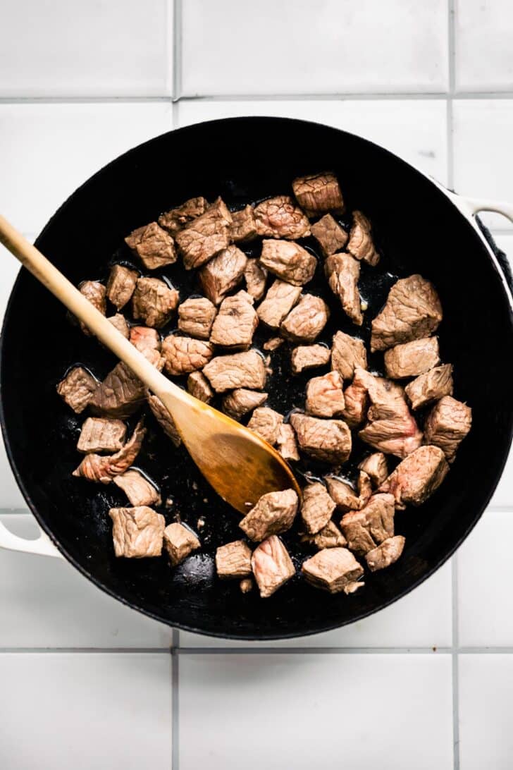 pieces of beef being browned in a skillet being stirred with a wooden spoon