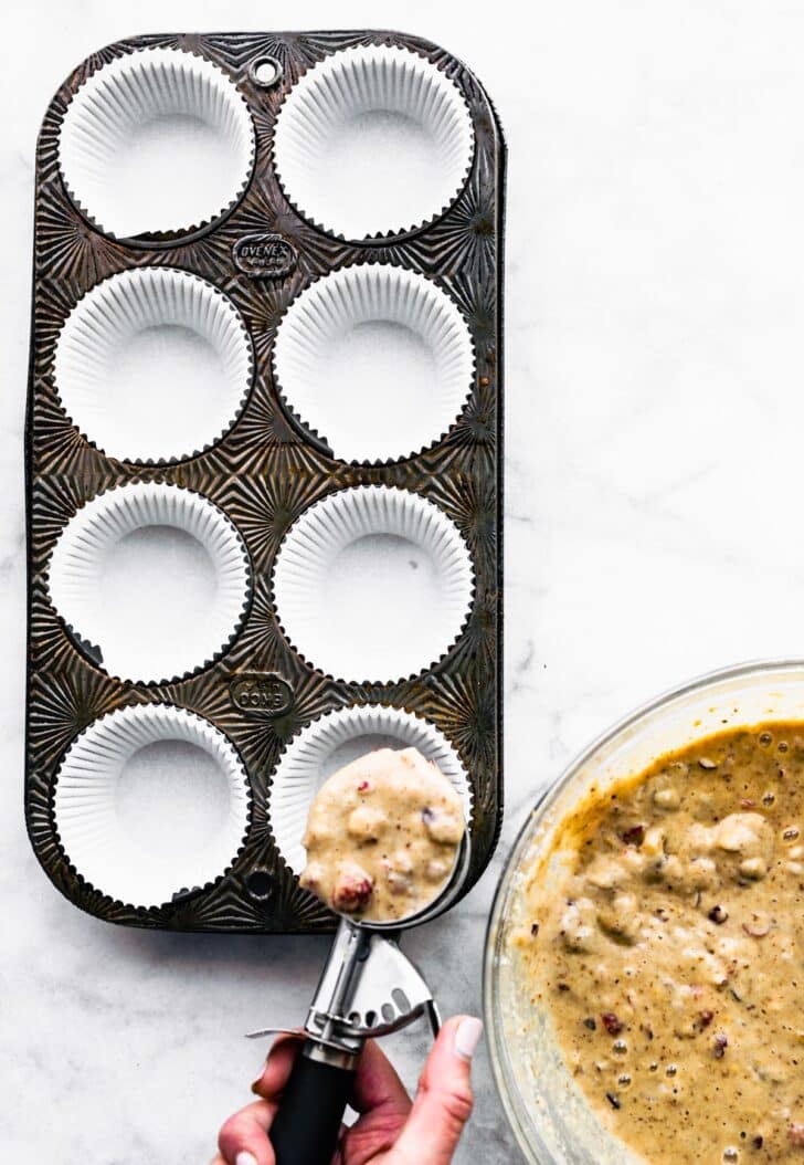 a muffin tin with liners and a hand holding a scoop of gluten free cranberry orange muffin batter