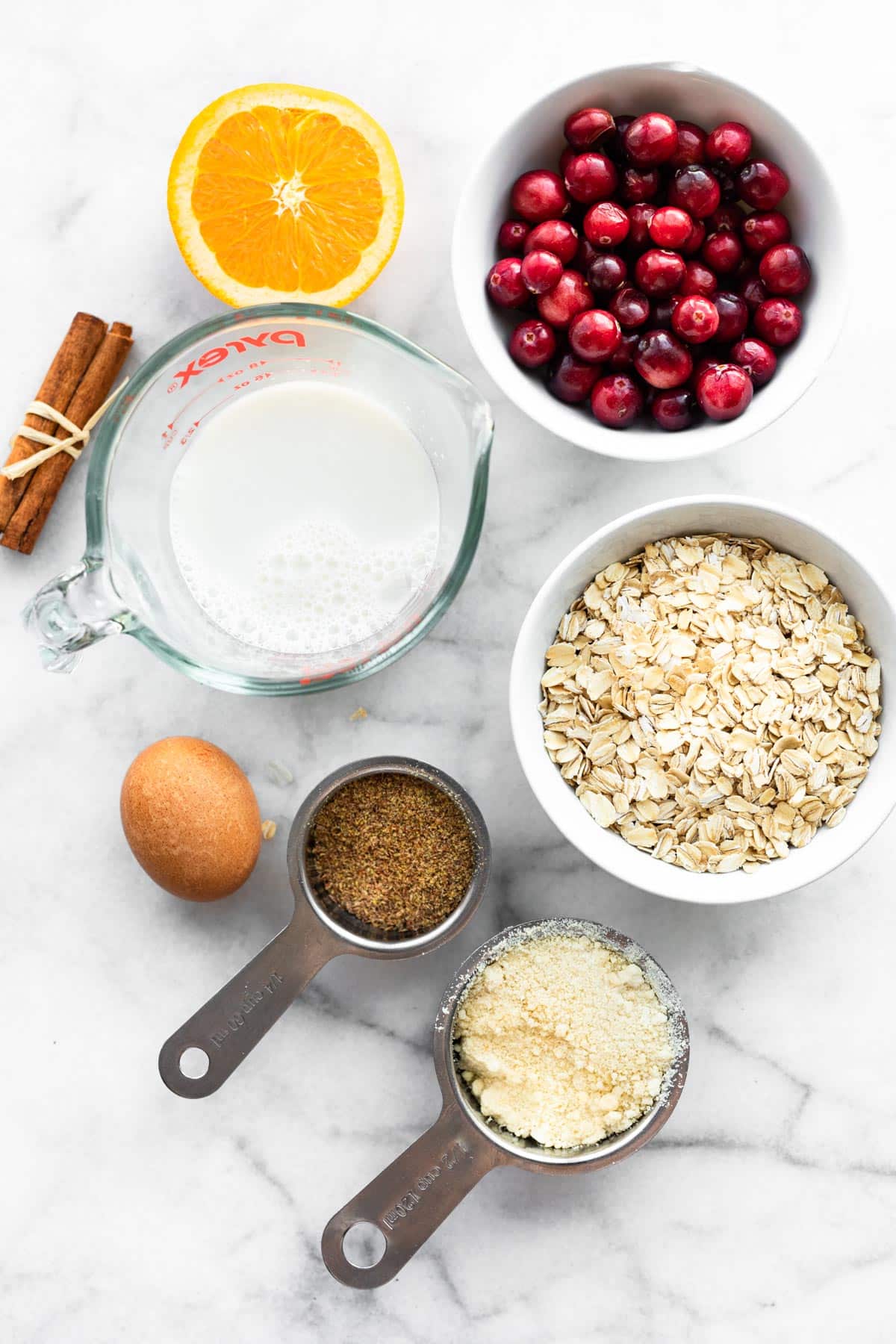 overhead ingredients including cranberries, orange, dairy-free milk, oats, an egg, and cinnamon sticks for gluten free cranberry orange muffins
