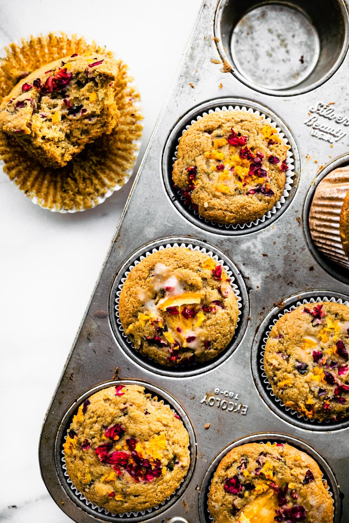 a muffin tin of gluten free cranberry orange muffins with one muffin on top of a liner to the side with a bite taken out