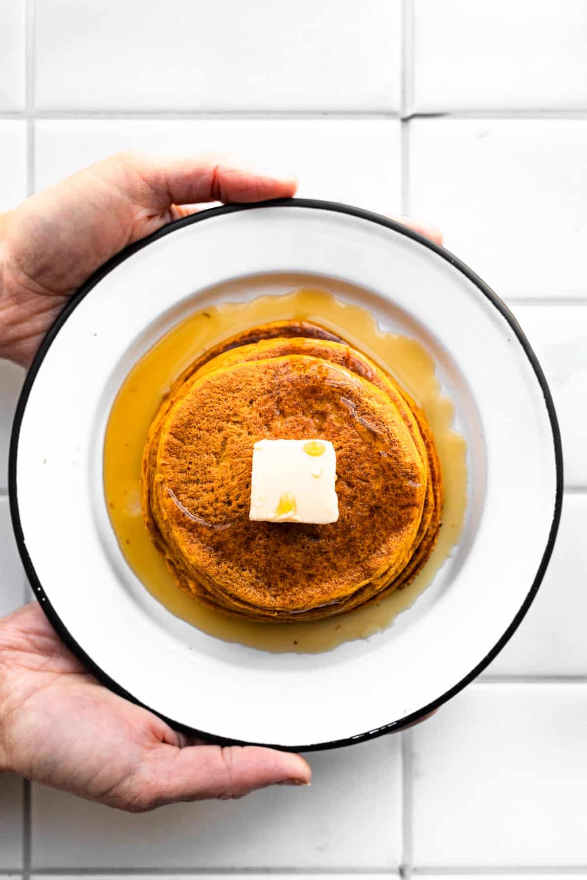 two hands holding a plate of vegan pumpkin pancakes topped with butter and syrup