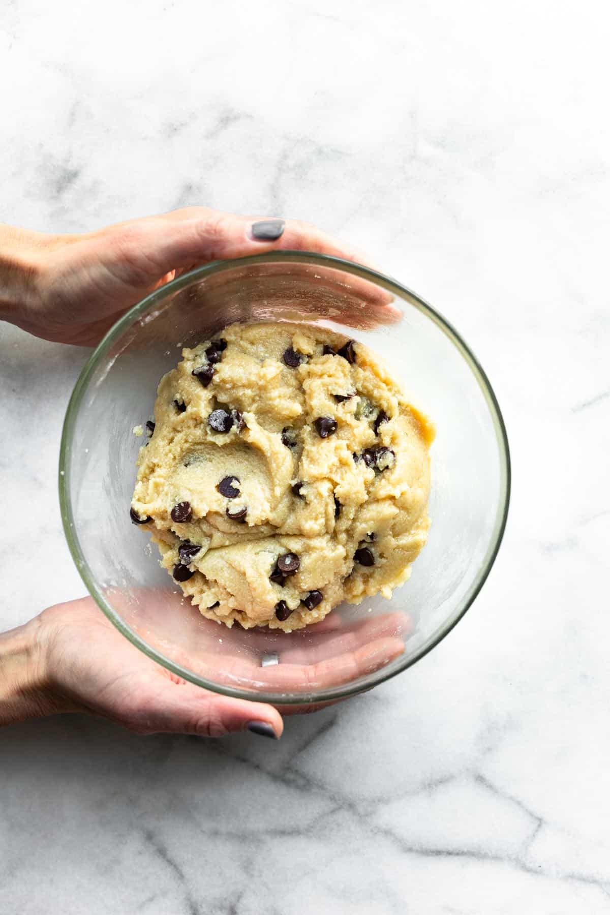 two hands holding a glass bowl full of vegan chocolate chip cookie dough