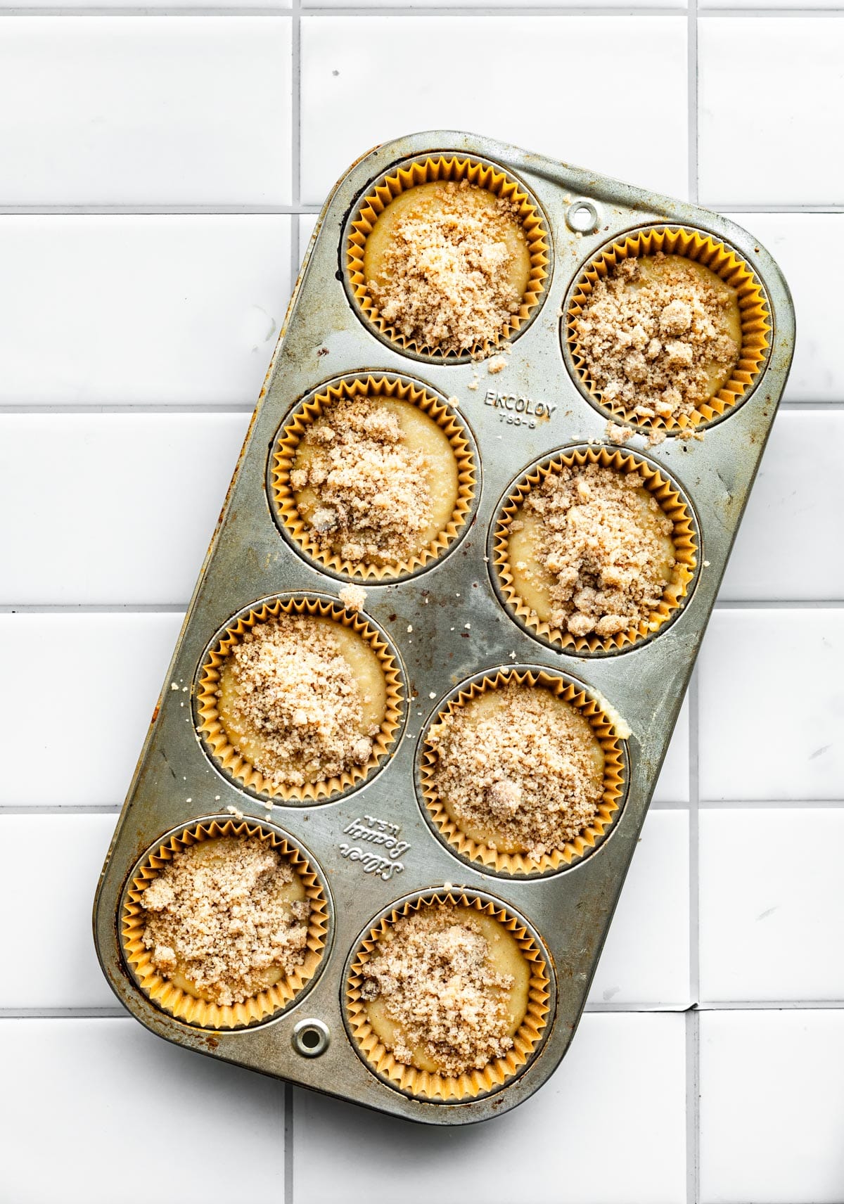 8 uncooked muffins in a muffin pan with crumb topping on a tile counter. 