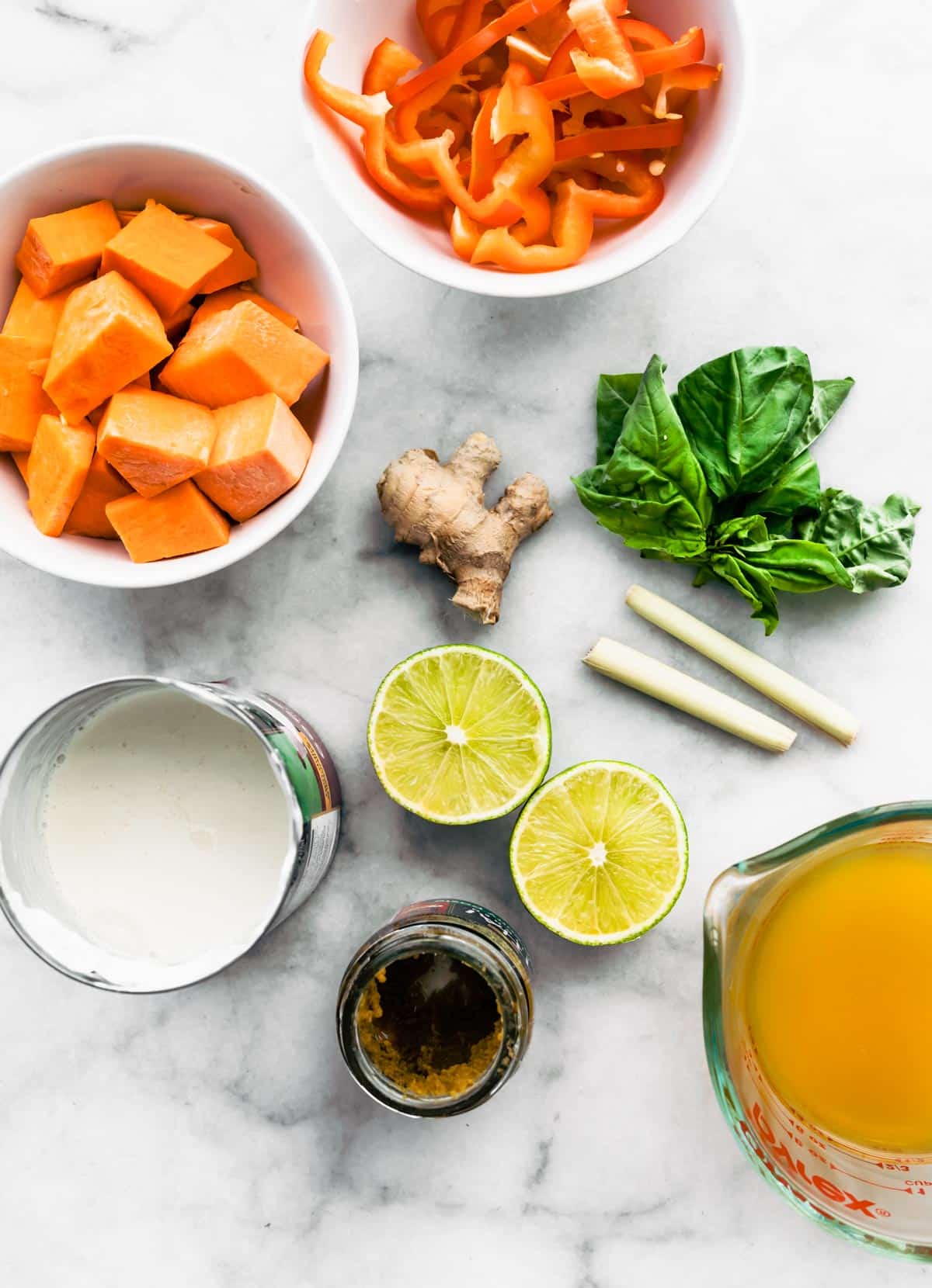 Overhead shot of ingredients on a counter top - lime split in half, ginger root, red peppers cut and in a bowl, sweet potatoes chopped up in a bowl, broth, coconut milk in a can and a few leaves of basil.