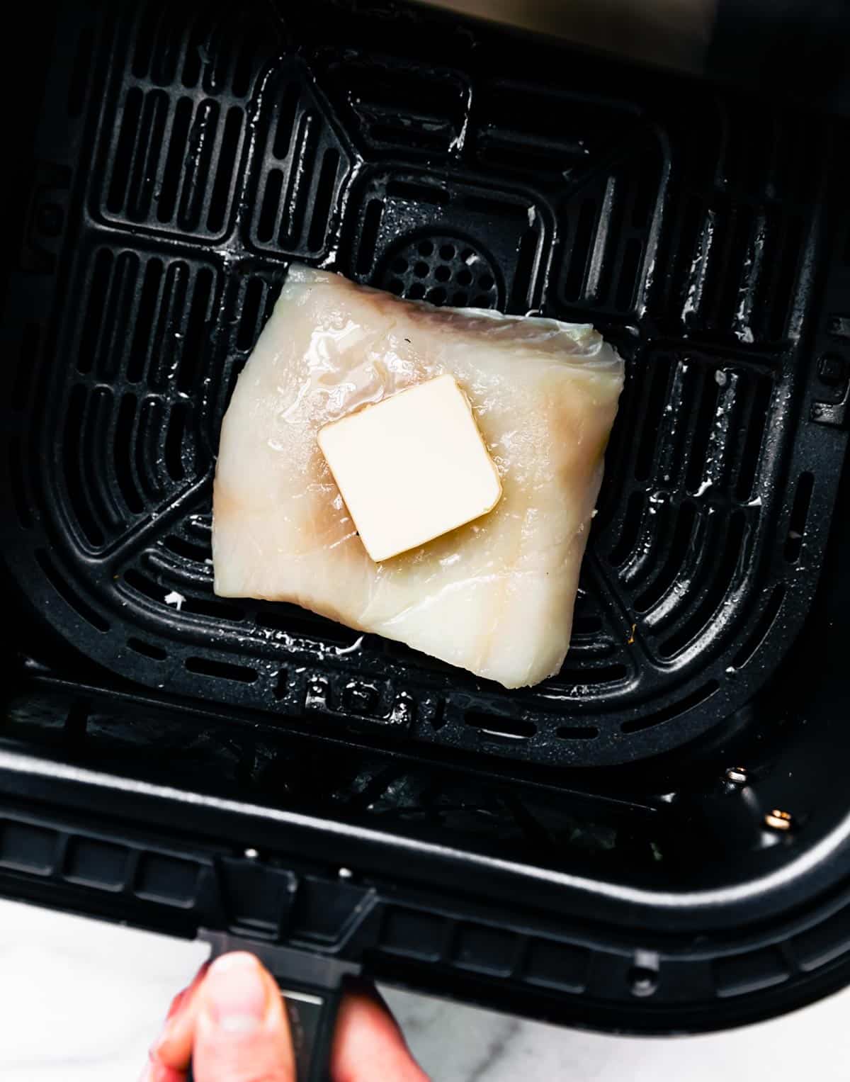 Overhead shot of a frozen fish fillet with a cube of butter on it sitting in an air fryer.